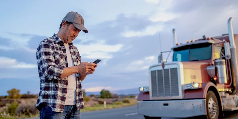 A truck driver standing in front of his truck, looking at his mobile phone.