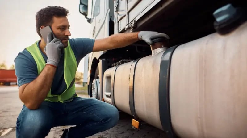 A truck driver holding a cell phone while inspecting the bottom side of his truck.